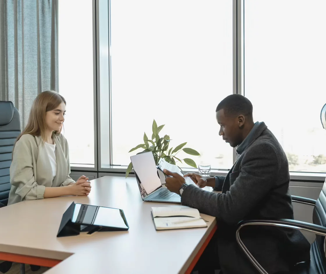A woman being interviewed by a man who is looking at a laptop screen and other documents