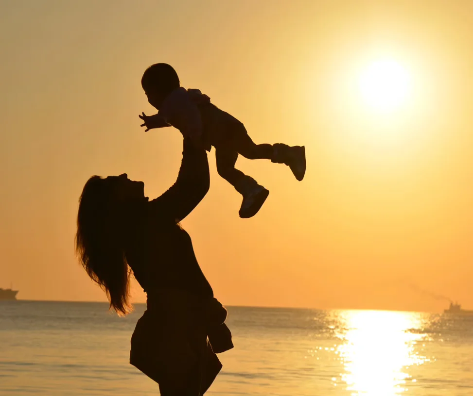 The silhoutte of a happy mother raising her baby in the air on a beach as the sun sets in the distance